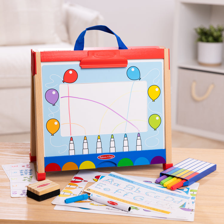 A playroom scene with The Melissa & Doug Take-Along Double-Sided Wooden Tabletop Activity Art Easel; Portable Travel Toy with Whiteboard, Chalkboard, Arts and Crafts Accessories