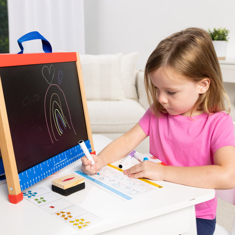 A kid playing with The Melissa & Doug Take-Along Double-Sided Wooden Tabletop Activity Art Easel; Portable Travel Toy with Whiteboard, Chalkboard, Arts and Crafts Accessories